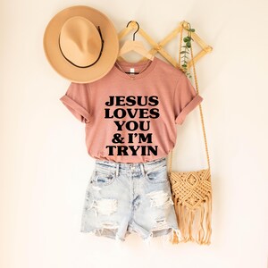 May include: A dusty rose t-shirt with the text "JESUS LOVES YOU & I'M TRYIN" in black block letters. The shirt is paired with denim shorts and a tan hat. A woven bag with fringe hangs nearby.