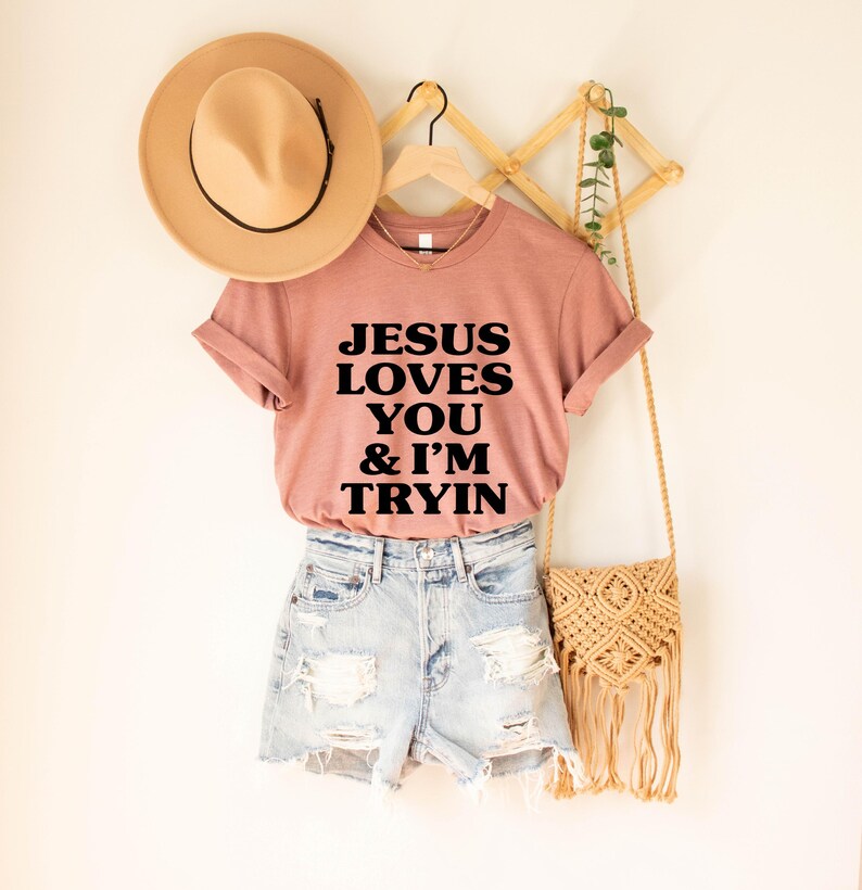 May include: A dusty rose t-shirt with the text "JESUS LOVES YOU & I'M TRYIN" in black block letters. The shirt is paired with denim shorts and a tan hat. A woven bag with fringe hangs nearby.