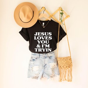May include: A black t-shirt with the text "JESUS LOVES YOU & I'M TRYIN" in white, paired with denim shorts, a tan hat, a macrame purse, and a gold necklace. The outfit is displayed on a white background.