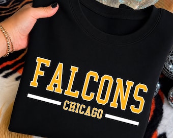 May include: A black crewneck sweatshirt featuring the word FALCONS in large yellow block letters above the word CHICAGO in smaller yellow letters, with a horizontal white line separating the two words and another white line below CHICAGO.