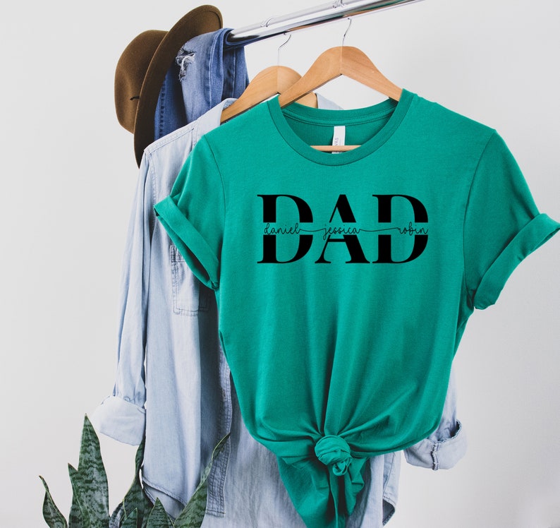 May include: Teal t-shirt with the word "DAD" in large black letters. The names "Daniel", "Jessica", and "Robin" are written in a smaller font size underneath the letters.