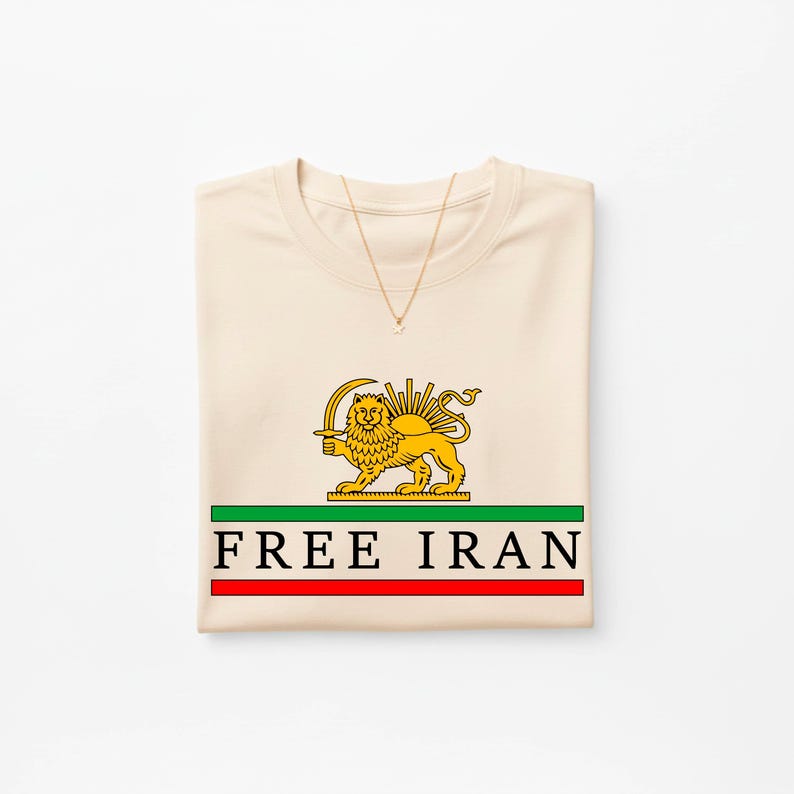 Może przedstawiać: Kremowa koszulka z złotym emblematem lwa z mieczem, promieniami słońca i napisem "FREE IRAN" w kolorze czarnym. Projekt jest umieszczony na zielonych i czerwonych paskach. Widoczny jest złoty naszyjnik.