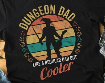 Peut inclure: T-shirt noir avec un design rétro. Le texte "DUNGEON DAD" est inscrit dans un cercle avec la silhouette d'un personnage tenant une épée et un bâton. Le texte "LIKE A REGULAR DAD BUT COOLER" est en dessous.