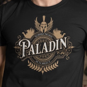 Op de afbeelding: Zwart T-shirt met een gouden afbeelding van een paladin-embleem. Het embleem toont een ridderhelm met vleugels, een schild en de tekst "To uphold the oath, or to break it". De tekst "Paladin" staat in een groot, sierlijk lettertype.