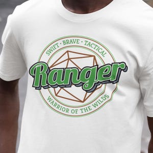 May include: White t-shirt featuring a green and black graphic with the word "Ranger" in a stylized font. The design includes a geometric shape and the text "Swift, Brave, Tactical" and "Warrior of the Wilds".