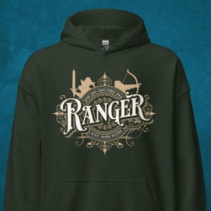 Puede incluir: Sudadera con capucha verde oscuro con un gráfico beige que dice "Ranger" en una fuente decorativa. El gráfico incluye siluetas de espada, arco y flecha, y otros elementos. También incluye el texto "Swiss Army Knife Forest Ninja Scout Sniper Soldier".