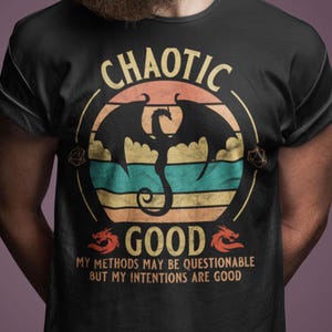 May include: Black t-shirt with a circular graphic featuring a dragon silhouette and the words "CHAOTIC GOOD." The design includes a sunset-colored background with horizontal stripes of orange, teal, and yellow. The text "MY METHODS MAY BE QUESTIONABLE BUT MY INTENTIONS ARE GOOD" is also included.