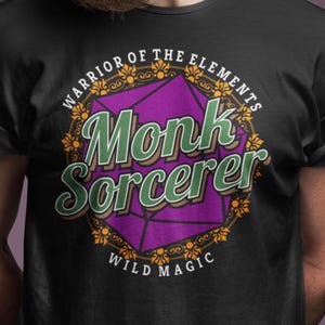Könnte beinhalten: Schwarzes T-Shirt mit einem lila und goldenen Design, das einen 20-seitigen Würfel mit dem Text "Warrior of the Elements Monk Sorcerer Wild Magic" zeigt.
