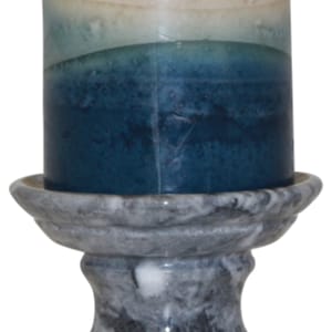May include: A lit, multi-toned candle in shades of cream, teal, and navy blue sits atop a gray marble candle holder. The candle holder has a decorative, ornate design. The candle is a pillar style.