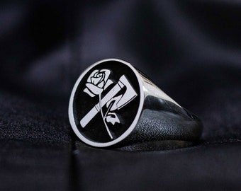 Può includere: Un anello d'argento con un design ovale nero che presenta una rosa bianca e un'ascia.