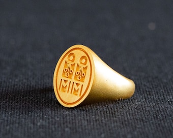 May include: A gold signet ring with a crest featuring the letters "MIM" and a design of three stylized flowers.