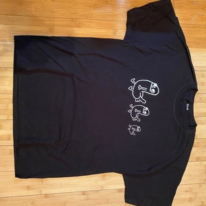 Pode incluir: Camiseta preta com um gráfico branco de três figuras de pau em linha, que ficam progressivamente menores. As figuras usam chapéus e têm um design simples e de desenho animado.