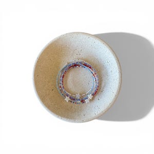 May include: A set of three circular beaded bracelets featuring small red, white, and blue glass beads. Each bracelet is accented with a single white star-shaped charm. The bracelets are arranged in a stack inside a round, textured beige stone bowl.