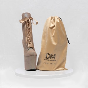 May include: A pair of light beige platform boots with a lace-up front and a satin ribbon. The boots are on a white pedestal and come with a beige dust bag that says "DM Dance Mistress pole shoes".