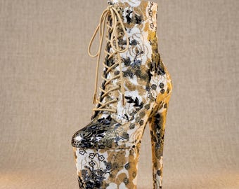 Op de afbeelding: Een enkelboot met hoge hak en vetersluiting aan de voorzijde met beige veters. De laars is bedekt met een stoffen patroon met een bloemendesign in wit, zwart en metallic goud. Het heeft een dikke plateauzool en een hoge, slanke stilettohak.