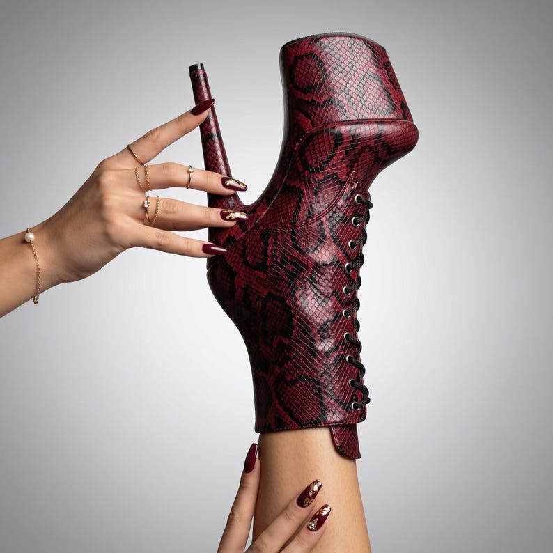 Op de afbeelding: Een enkellaars met hoge hak en een donkerrood en zwart slangenleermotief. De laars heeft een dikke plateauzool, een zeer hoge stilettohak en een vetersluiting aan de voorzijde met zwarte veters en zilverkleurige metalen oogjes.