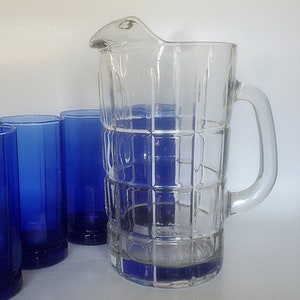 May include: A set of clear glass pitcher and blue glass tumblers. The pitcher has a square pattern and a handle. The tumblers have a ribbed design.