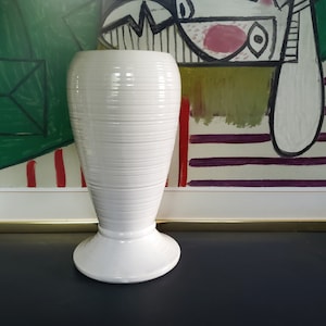 Könnte beinhalten: Weiße Keramikvase mit geripptem Design und breiter Basis. Die Vase steht auf einer dunklen Oberfläche.