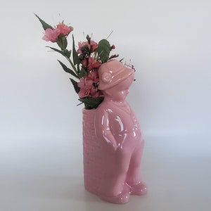 May include: Pink ceramic vase shaped like a boy wearing a hat. The vase has a brick pattern on the back and is filled with pink flowers.