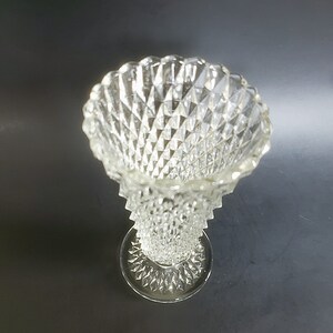 May include: Clear glass vase with a diamond pattern. The vase has a wide, scalloped rim and a pedestal base. The glass is transparent, allowing light to pass through. The vase is likely used for decorative purposes, such as holding flowers.