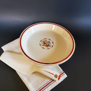 May include: A white pie plate with a red rim and a floral design in the center. The plate is sitting on a white cloth with red and blue stripes.