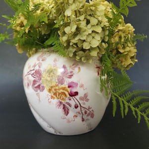 Könnte beinhalten: Eine weiße Keramikvase mit einem Blumendesign in Gelb, Rosa und Burgunder. Die Vase ist mit getrockneten gelben Blumen und grünen Farnblättern gefüllt. Die Vase hat eine einzigartige, geschwungene Form.