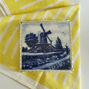 May include: A blue and white ceramic tile with a silver frame depicting a traditional Dutch windmill with a house and trees. The tile has a blue and white design.