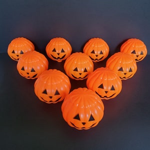 May include: A collection of ten bright orange plastic jack-o'-lantern buckets arranged in a triangular formation. Each pumpkin-shaped container features a molded ribbed texture and a black painted face with triangular eyes, a nose, and a grin.