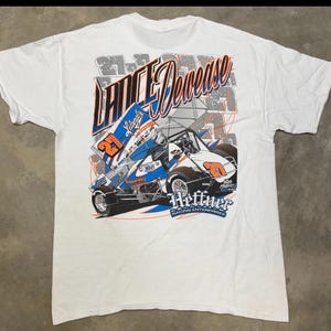 May include: White t-shirt featuring a graphic of a sprint car with the text "Lance Dewease" in orange and blue. The car is blue, white, and orange with the number 21. The shirt also has the text "Heffner Racing Enterprises".