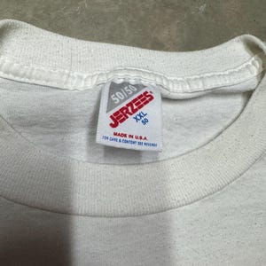 Puede incluir: Primer plano de un cuello de camiseta blanca con una etiqueta Jerzees. La etiqueta dice "50/50" y "XXL 50" con el texto "MADE IN U.S.A." y "FOR CARE & CONTENT SEE REVERSE". La camiseta tiene un patr&oacute;n de cuadros en la parte inferior.