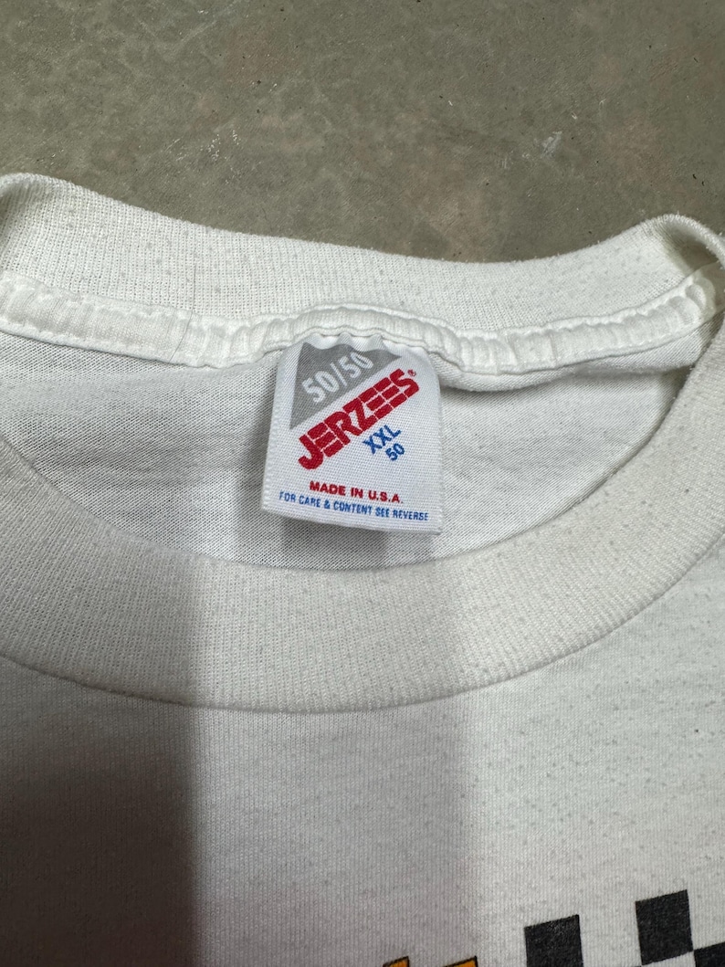 Puede incluir: Primer plano de un cuello de camiseta blanca con una etiqueta Jerzees. La etiqueta dice "50/50" y "XXL 50" con el texto "MADE IN U.S.A." y "FOR CARE & CONTENT SEE REVERSE". La camiseta tiene un patr&oacute;n de cuadros en la parte inferior.