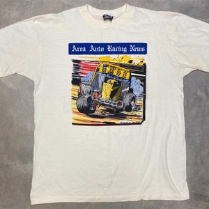 May include: White t-shirt with a graphic of a yellow race car on a dirt track. The graphic is surrounded by a blue border and the text "Area Auto Racing News".