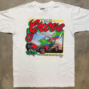 Puede incluir: Camiseta blanca con un gráfico colorido de un coche de carreras y el texto "THE GROVE" en rojo y verde. La camiseta también muestra "COTTAGE GROVE, OR" y los nombres "Kris Dickinson" y "Mark Dziadzioz".