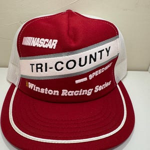 May include: A red and white NASCAR trucker hat with the text "TRI-COUNTY SPEEDWAY" and "Winston Racing Series". The hat has a white mesh back and a curved brim with white trim. The word "NASCAR" is printed on the front.