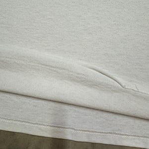 Puede incluir: Primer plano de una camiseta blanca, que muestra la textura de la tela y el dobladillo cosido. La camiseta parece estar hecha de un material suave, posiblemente desgastado. La imagen se centra en los detalles de la prenda.