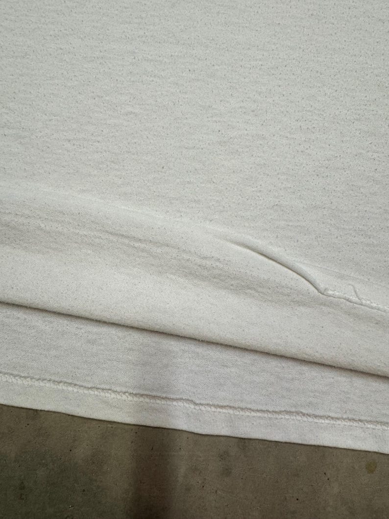 Puede incluir: Primer plano de una camiseta blanca, que muestra la textura de la tela y el dobladillo cosido. La camiseta parece estar hecha de un material suave, posiblemente desgastado. La imagen se centra en los detalles de la prenda.