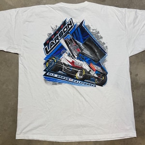 May include: White t-shirt featuring a graphic of a race car with the name "Kyle Larson" and the text "Elk Grove California". The design incorporates blue, red, and black colors, with a racing theme.