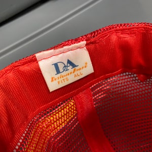 May include: Red mesh baseball cap with a white label that reads "DA Designer Apparel FITS ALL".