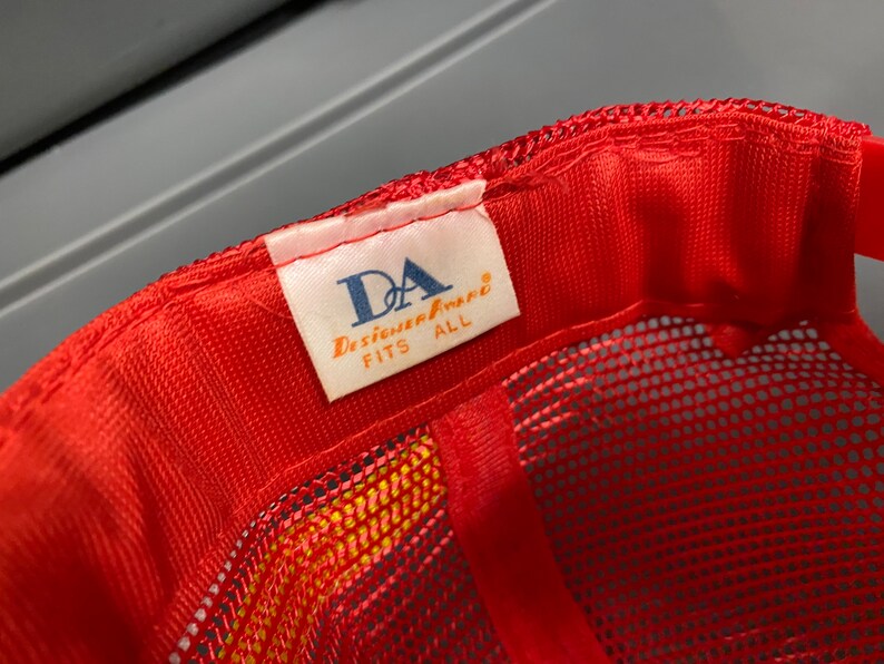 May include: Red mesh baseball cap with a white label that reads "DA Designer Apparel FITS ALL".