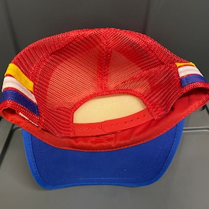 May include: Red and blue mesh trucker hat with a yellow and white stripe. The hat has a plastic adjustable strap in the back.