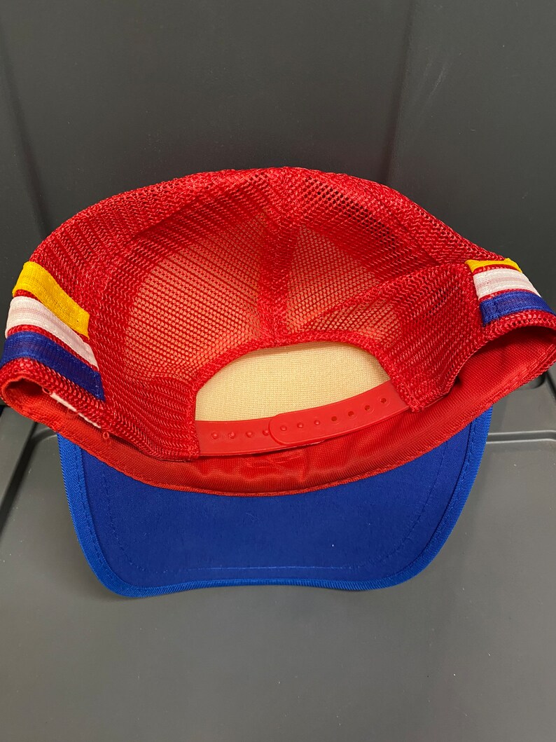 May include: Red and blue mesh trucker hat with a yellow and white stripe. The hat has a plastic adjustable strap in the back.