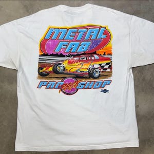May include: White t-shirt featuring a colorful graphic of a race car with the text "METAL FAB PRO SHOP". The design includes a checkered flag and a sunset background. The shirt is a classic crew neck.