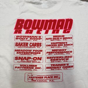 Puede incluir: Camiseta blanca con un gr&aacute;fico rojo y rosa que dice "BOWMAN RACING". Debajo, varios logotipos rectangulares rojos con texto blanco, incluyendo "BAKER CARBS" y "MIDWAY PERFORMANCE".