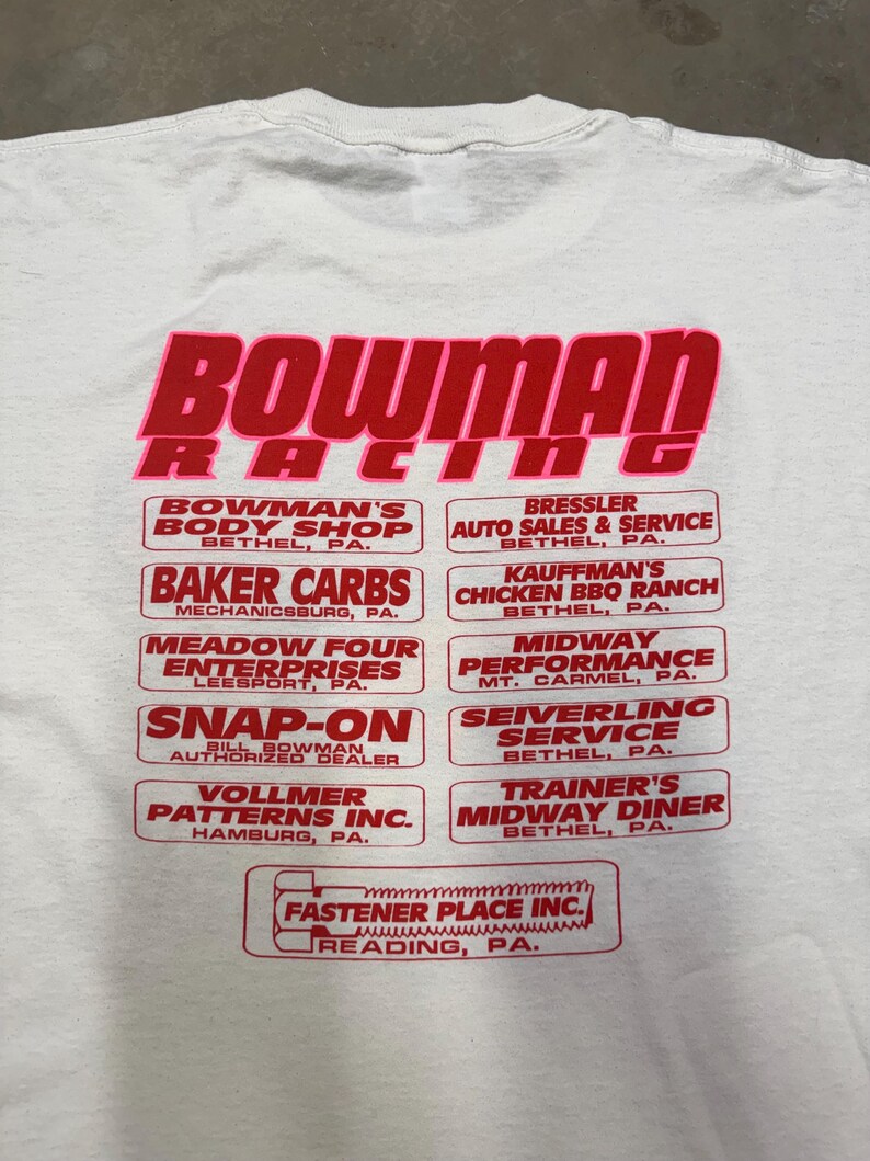 Puede incluir: Camiseta blanca con un gr&aacute;fico rojo y rosa que dice "BOWMAN RACING". Debajo, varios logotipos rectangulares rojos con texto blanco, incluyendo "BAKER CARBS" y "MIDWAY PERFORMANCE".