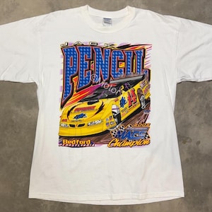 Puede incluir: Camiseta blanca con un coche de carreras amarillo. El diseño incluye el texto "JACK PENCIL" en azul y rojo, junto con "Bedford Pennsylvania" y "2002 Mags Champion".