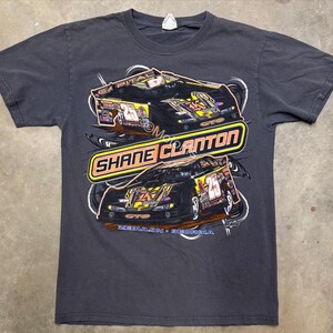 May include: Dark gray t-shirt featuring a graphic of two race cars with the text "SHANE CLANTON" in a yellow and orange design. The shirt also includes the text "ZEBULON, GEORGIA".