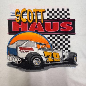 Puede incluir: Camiseta blanca con un gr&aacute;fico de coche de carreras. El dise&ntilde;o incluye el texto "SCOTT HAUS" en naranja y amarillo, y un coche de carreras blanco con detalles en naranja y azul. Se muestra el n&uacute;mero "70". Un dise&ntilde;o de bandera a cuadros est&aacute; en el fondo.