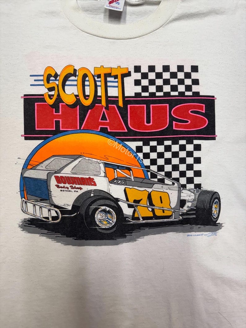 Puede incluir: Camiseta blanca con un gr&aacute;fico de coche de carreras. El dise&ntilde;o incluye el texto "SCOTT HAUS" en naranja y amarillo, y un coche de carreras blanco con detalles en naranja y azul. Se muestra el n&uacute;mero "70". Un dise&ntilde;o de bandera a cuadros est&aacute; en el fondo.