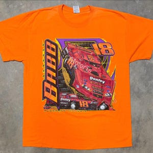 May include: Bright orange t-shirt featuring a graphic of a red race car with the number 18. The design includes the name "BABB" vertically and the text "Donley Auto Parts". The shirt is made of a soft material.