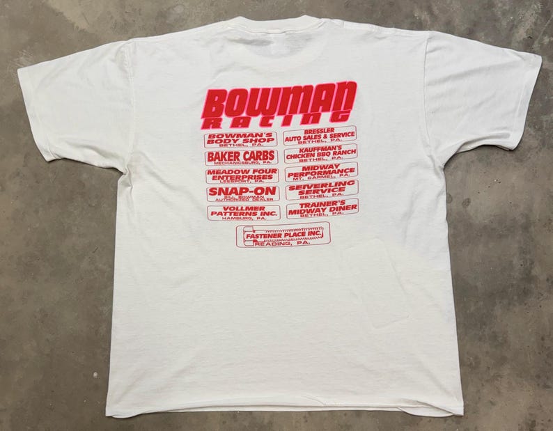 Puede incluir: Camiseta blanca con el texto rojo "BOWMAN RACING" y varios nombres de empresas. La camiseta tiene mangas cortas y cuello redondo. La camiseta est&aacute; hecha de un material suave y c&oacute;modo.